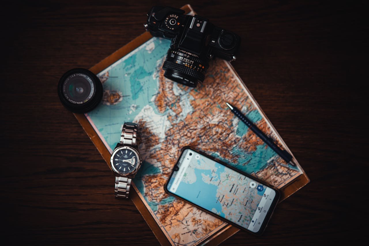 Top view of travel essentials including a map, camera, phone, watch, and lens on a wooden surface.